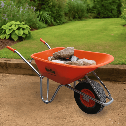 Belle Warrior wheelbarrow with puncture-proof tyre, filled with rubble and positioned on a compacted soil surface in a landscaped garden. The wheelbarrow features an orange HDPE tray and ergonomic handles with greenery and lawn in the background
